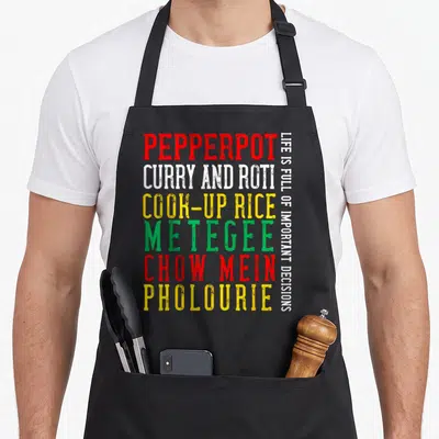 Guyana food aprons - Stacked text listing pepperpot, curry, roti, and other dishes in red, white, yellow, and green — shown on a black cotton canvas apron against a white background