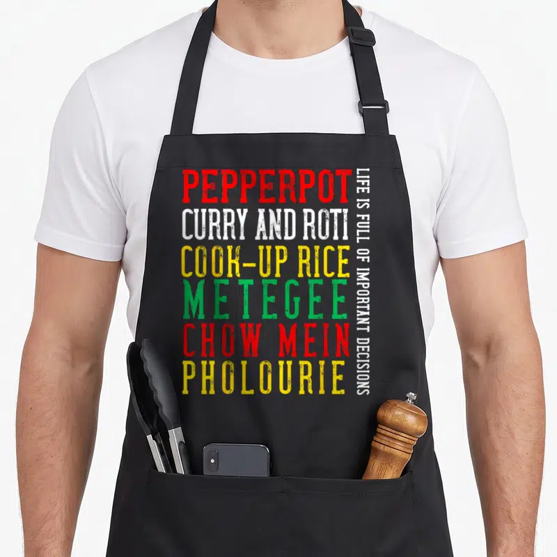 Guyana food aprons - Stacked text listing pepperpot, curry, roti, and other dishes in red, white, yellow, and green — shown on a black cotton canvas apron against a white background