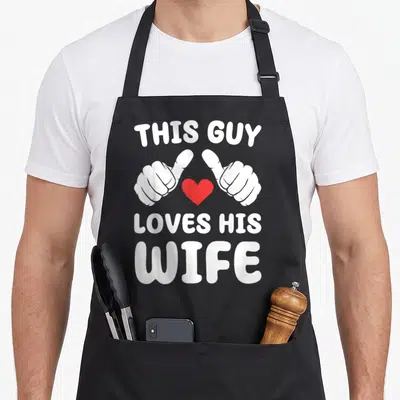 Wife valentine's day apron - Cartoon hands pointing at red heart with white text THIS GUY LOVES HIS WIFE — shown on a black cotton canvas apron against a white background