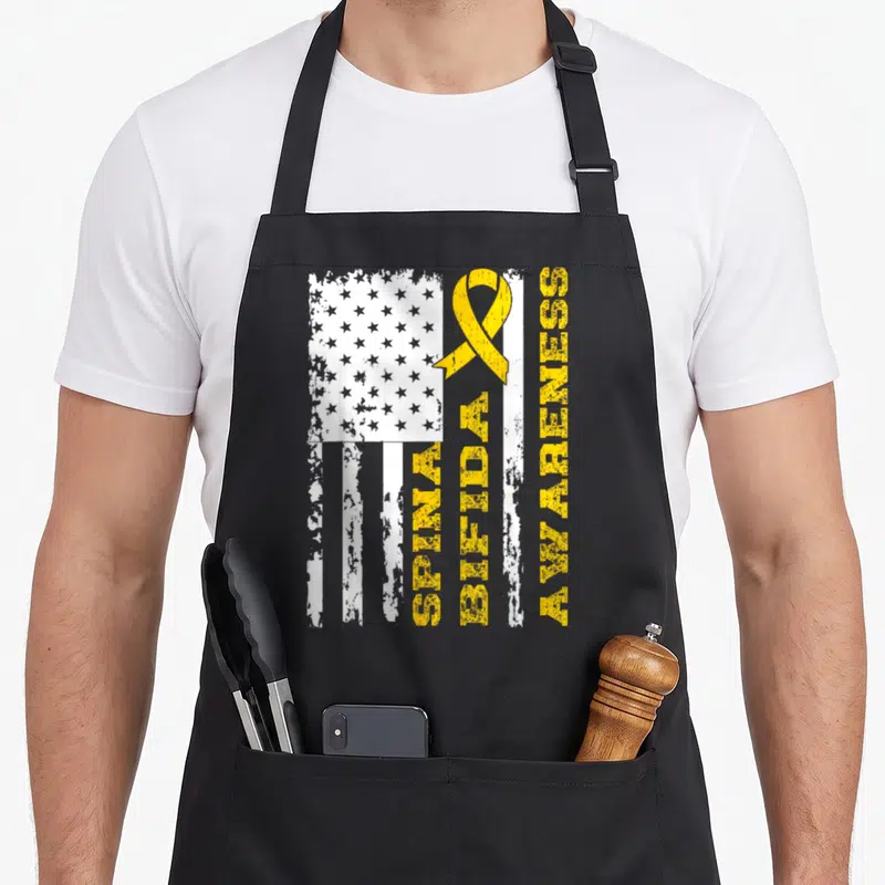 Bifida awareness ribbon apron - Distressed white flag with yellow ribbon and vertical text SPINA BIFIDA AWARENESS — shown on a black cotton canvas apron against a white background