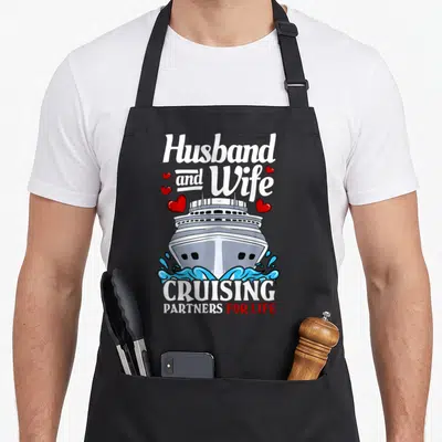 Cruising husband wife aprons - Cartoon cruise ship with red hearts and partners for life text — shown on a black cotton canvas apron against a white background