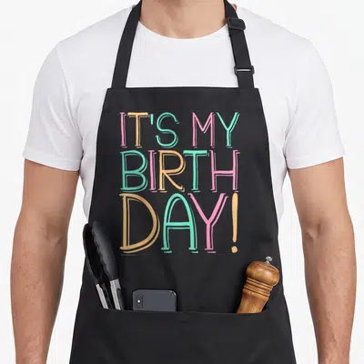Pastel birthday apron - Stacked IT'S MY BIRTH DAY text in pink orange teal hand-drawn font — shown on a black cotton canvas apron against a white background