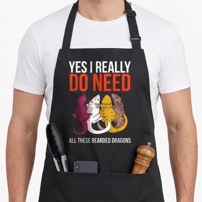 Bearded do i need an apron - Four cartoon bearded dragons in purple, yellow, white, red, and brown clustered under bold — shown on a black cotton canvas apron against a white background