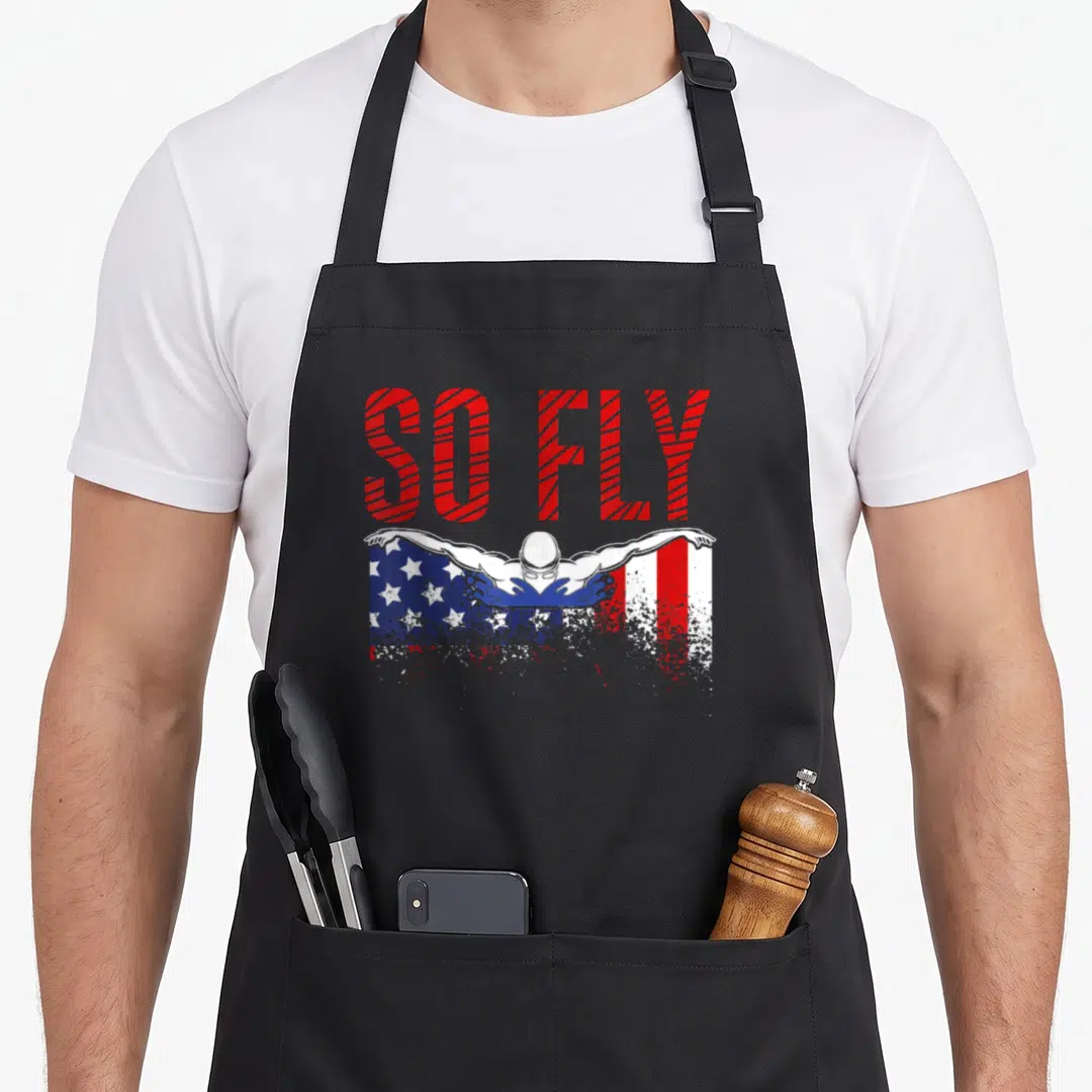 Fly sports team aprons - SO FLY text above swimmer on distressed red white blue USA flag background — shown on a black cotton canvas apron against a white background