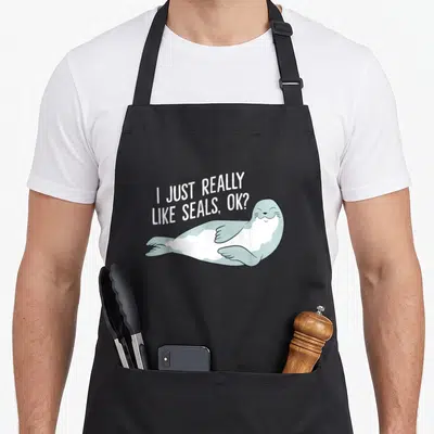 Funny seal apron - Cartoon seal lying down with white text I just really like seals ok — shown on a black cotton canvas apron against a white background