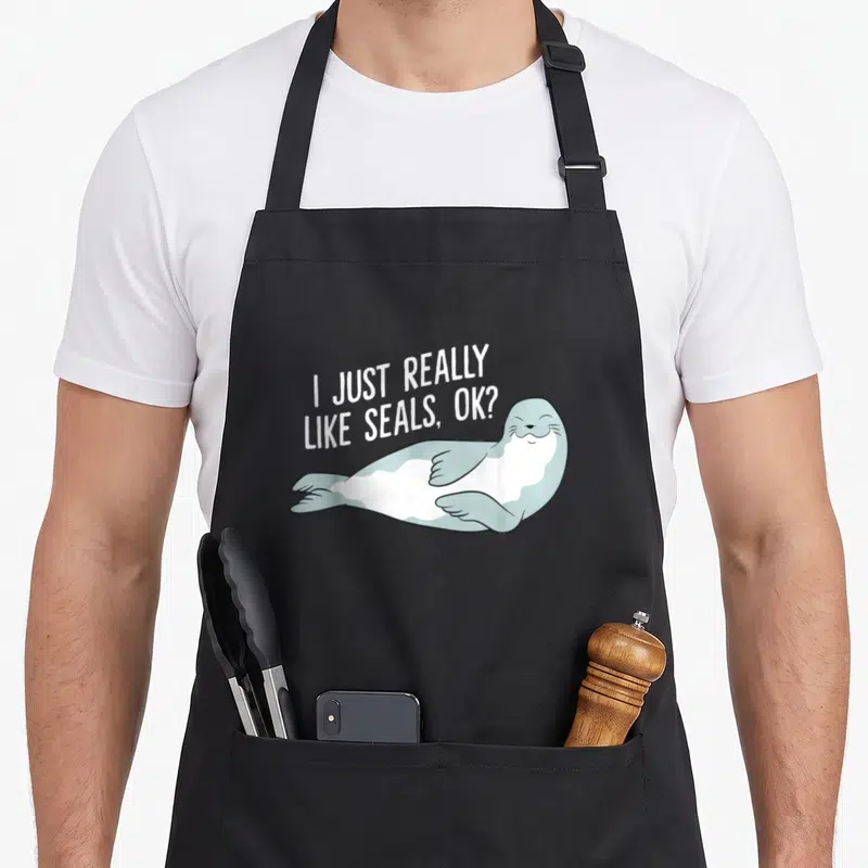 Funny seal apron - Cartoon seal lying down with white text I just really like seals ok — shown on a black cotton canvas apron against a white background