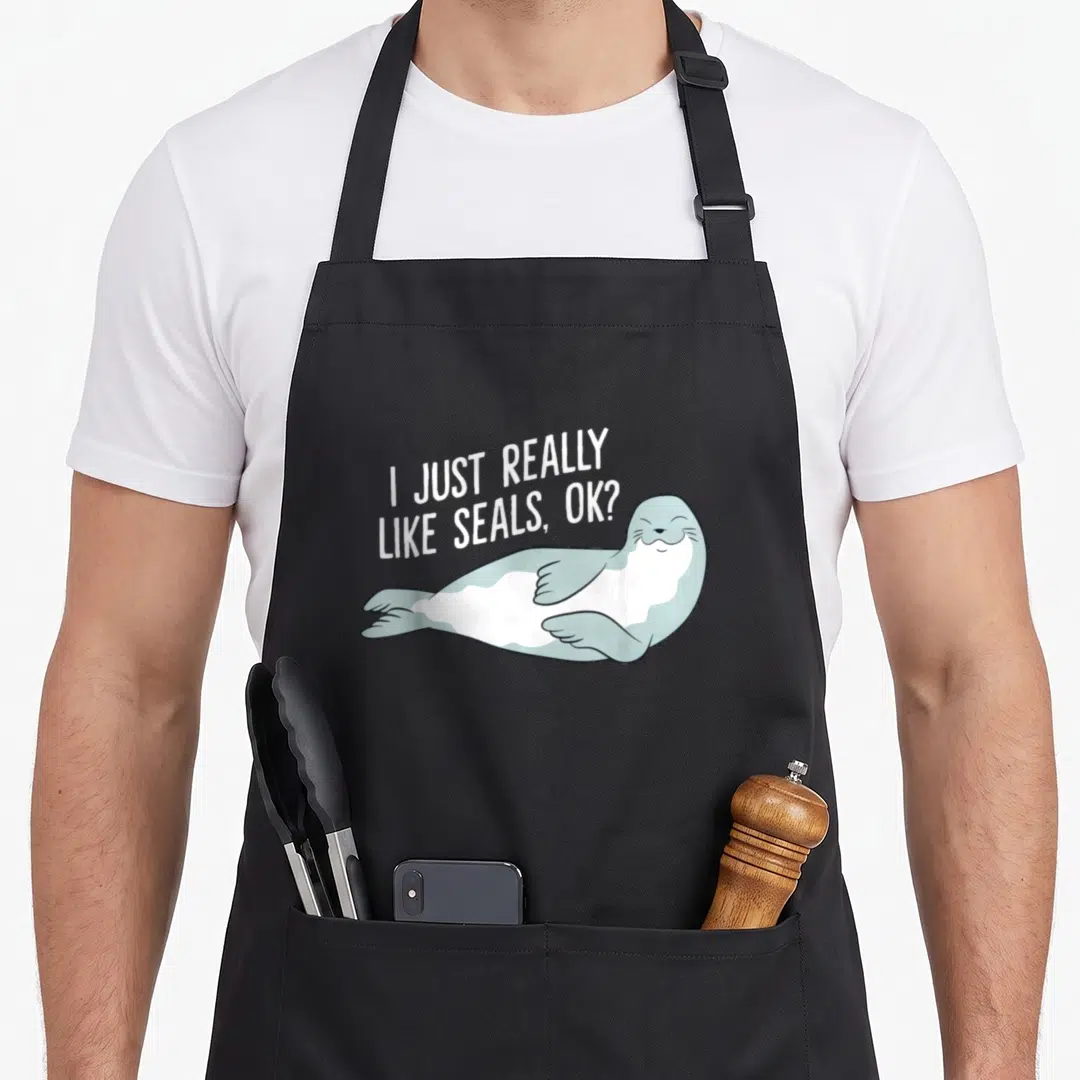 Funny seal apron - Cartoon seal lying down with white text I just really like seals ok — shown on a black cotton canvas apron against a white background