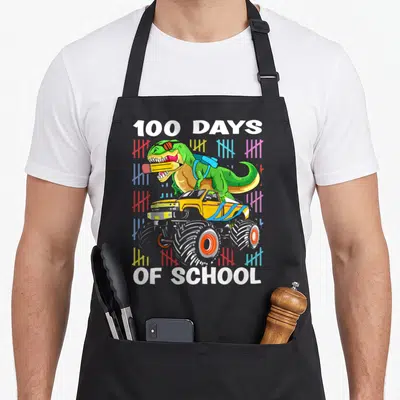100th school truck apron - T-Rex on monster truck with 100 DAYS OF SCHOOL text and colorful tally marks — shown on a black cotton canvas apron against a white background