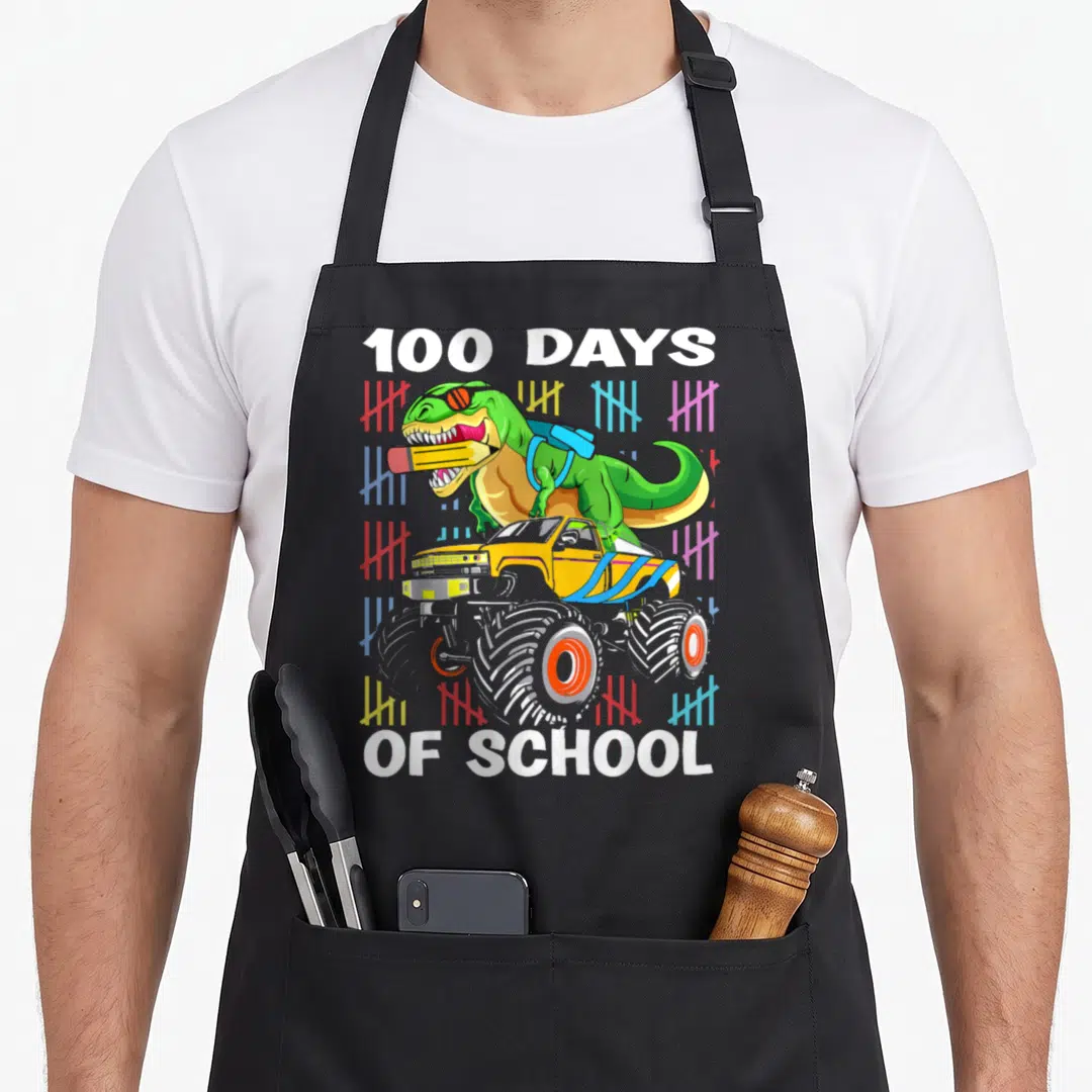 100th school truck apron - T-Rex on monster truck with 100 DAYS OF SCHOOL text and colorful tally marks — shown on a black cotton canvas apron against a white background