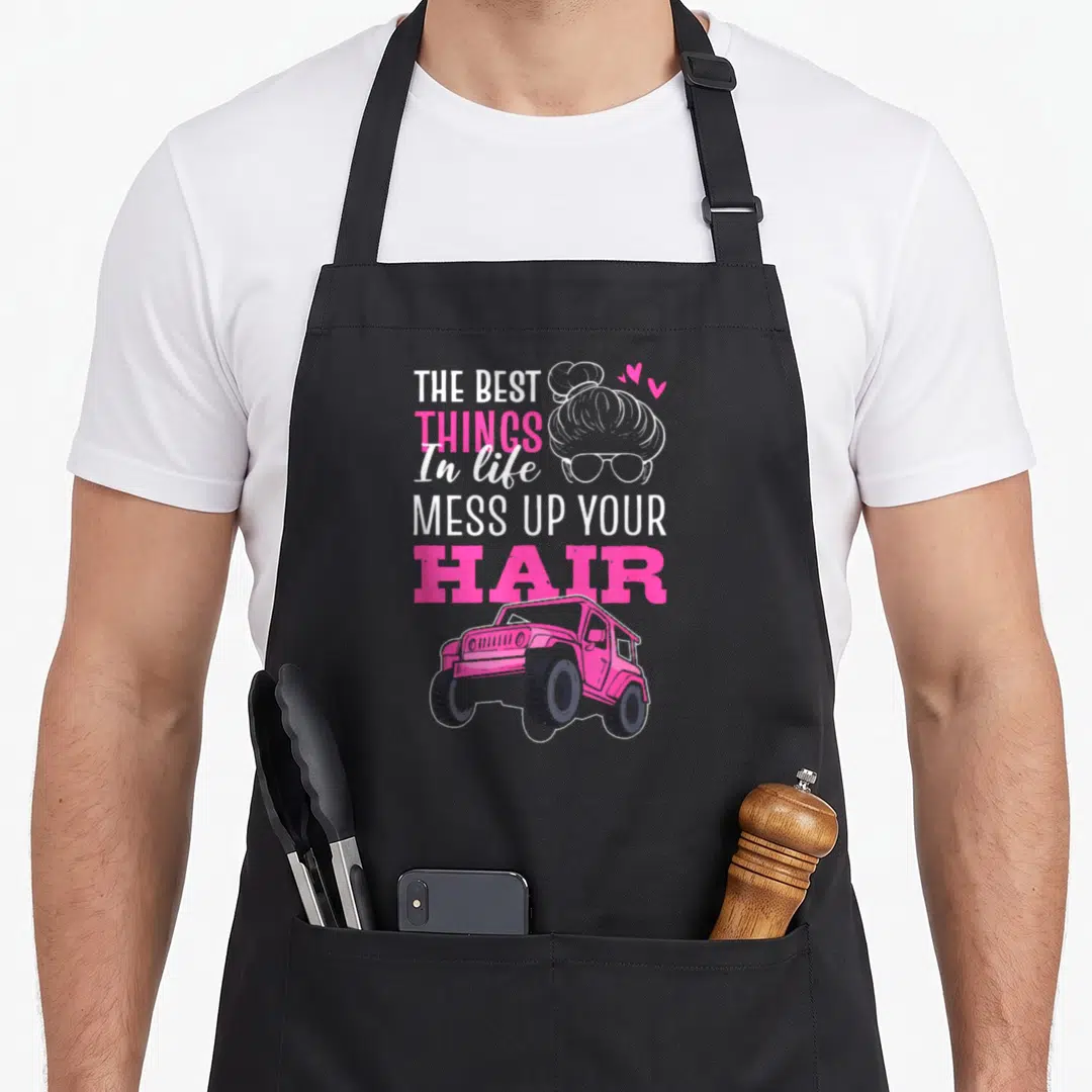 Mess hair apron - Pink jeep and hair bun with sunglasses below stacked text reading The Best Things Mess Up Your Hair — shown on a black cotton canvas apron against a white background