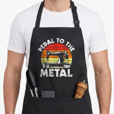 Pedal machine apron - PEDAL TO THE METAL text arching over a sewing machine and accessories on a striped sunset — shown on a black cotton canvas apron against a white background