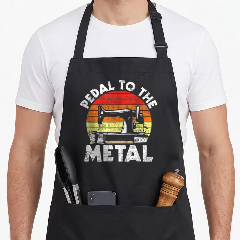 Pedal machine apron - PEDAL TO THE METAL text arching over a sewing machine and accessories on a striped sunset — shown on a black cotton canvas apron against a white background