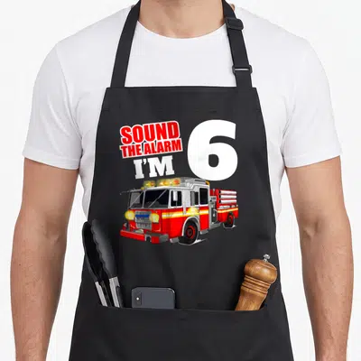 Six truck apron - SOUND THE ALARM I'M 6 text with red fire truck illustration — shown on a black cotton canvas apron against a white background