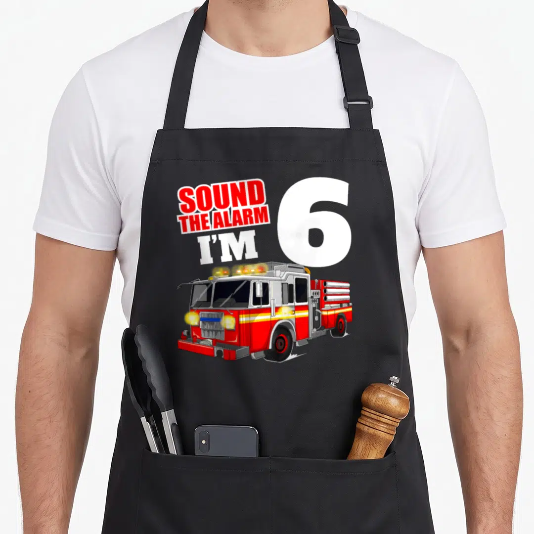 Six truck apron - SOUND THE ALARM I'M 6 text with red fire truck illustration — shown on a black cotton canvas apron against a white background