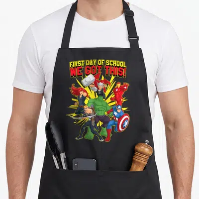 Funny school novelty apron - Avengers cluster below FIRST DAY OF SCHOOL WE GOT THIS! text in cartoon style with yellow — shown on a black cotton canvas apron against a white background