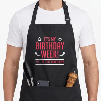 Funny whole birthday apron - Stacked pink text IT'S MY BIRTHDAY WEEK flanked by white stars and horizontal lines — shown on a black cotton canvas apron against a white background