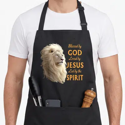 Loved christian apron - Realistic lion head beside stacked orange text reading Blessed by God Loved by Jesus Led — shown on a black cotton canvas apron against a white background