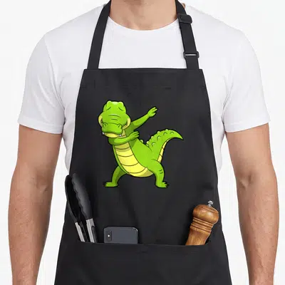 Dabbing tap dancing apron - Green cartoon crocodile performing a dab pose with a yellow segmented belly — shown on a black cotton canvas apron against a white background