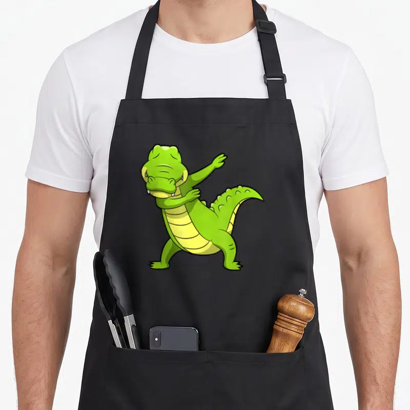 Dabbing tap dancing apron - Green cartoon crocodile performing a dab pose with a yellow segmented belly — shown on a black cotton canvas apron against a white background