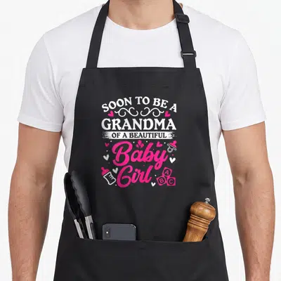 Soon grandma apron - Stacked white and pink text reading SOON TO BE A GRANDMA OF A BEAUTIFUL Baby Girl with icons — shown on a black cotton canvas apron against a white background