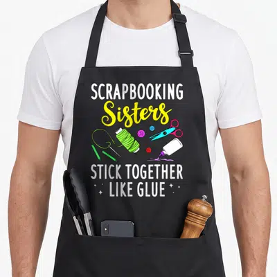 Scrapbooking craft apron - Scrapbooking Sisters Stick Together Like Glue text with cartoon scissors, thread, buttons — shown on a black cotton canvas apron against a white background