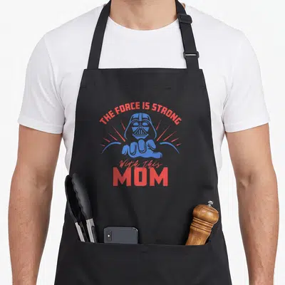 Force strong mother's day aprons - Darth Vader head and hand in blue with red rays and text THE FORCE IS STRONG — shown on a black cotton canvas apron against a white background
