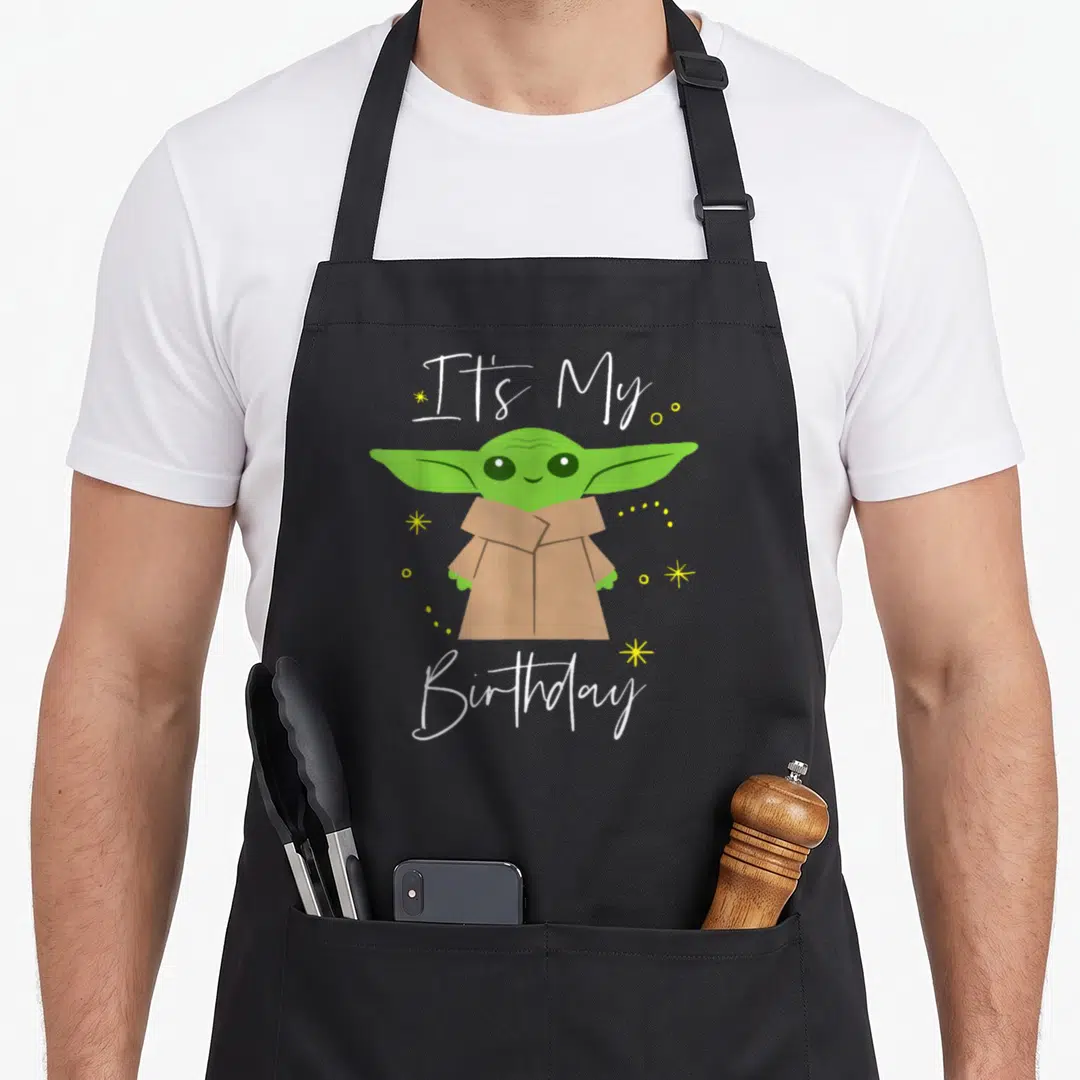 Cool age birthday apron - Cartoon Grogu with It's My Birthday text surrounded by yellow stars and dots — shown on a black cotton canvas apron against a white background