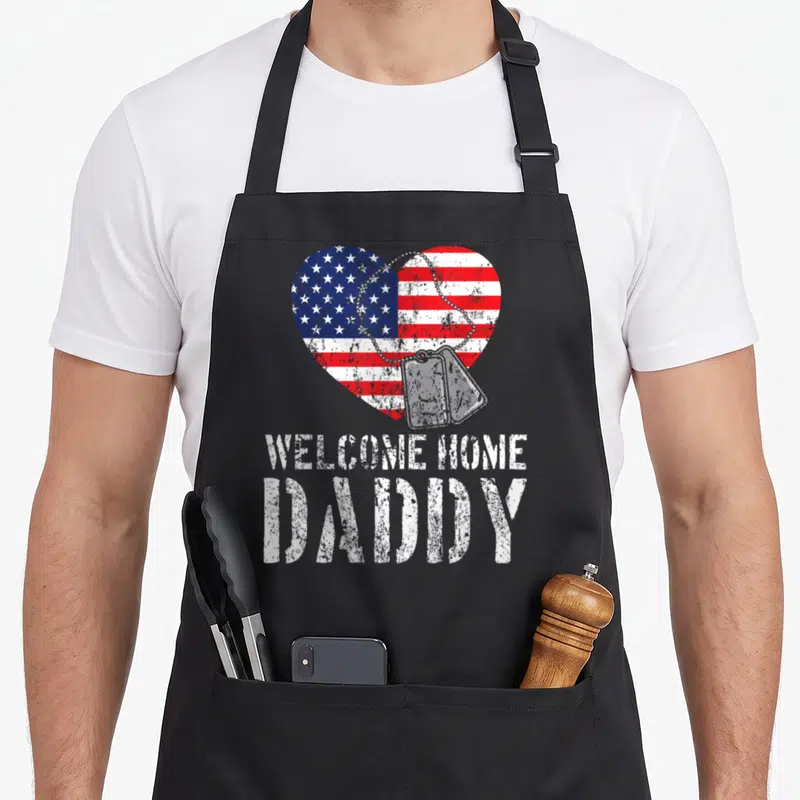 Welcome daddy apron - Distressed heart with flag and dog tags saying Welcome Home Daddy — shown on a black cotton canvas apron against a white background