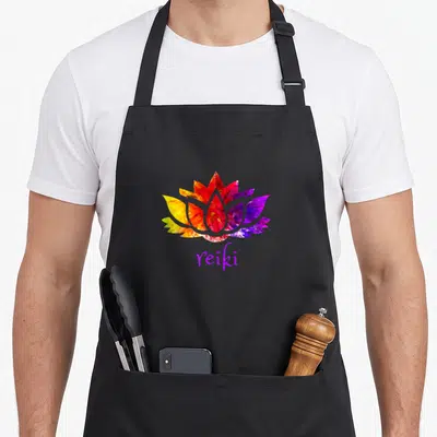 Reiki energy apron - Faceted lotus flower in rainbow colors above purple reiki text on black background — shown on a black cotton canvas apron against a white background