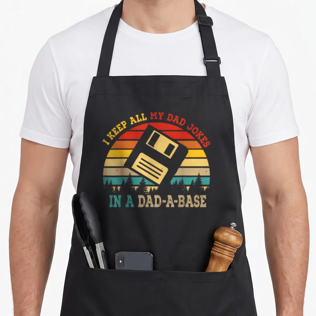 Vintage base dad joke apron - Retro sunset with grey floppy disk and text I KEEP ALL MY DAD JOKES IN A DAD-A-BASE — shown on a black cotton canvas apron against a white background