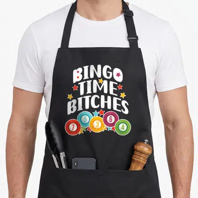 Cute player bingo apron - BINGO TIME BITCHES text surrounded by colorful stars and five numbered bingo chips — shown on a black cotton canvas apron against a white background