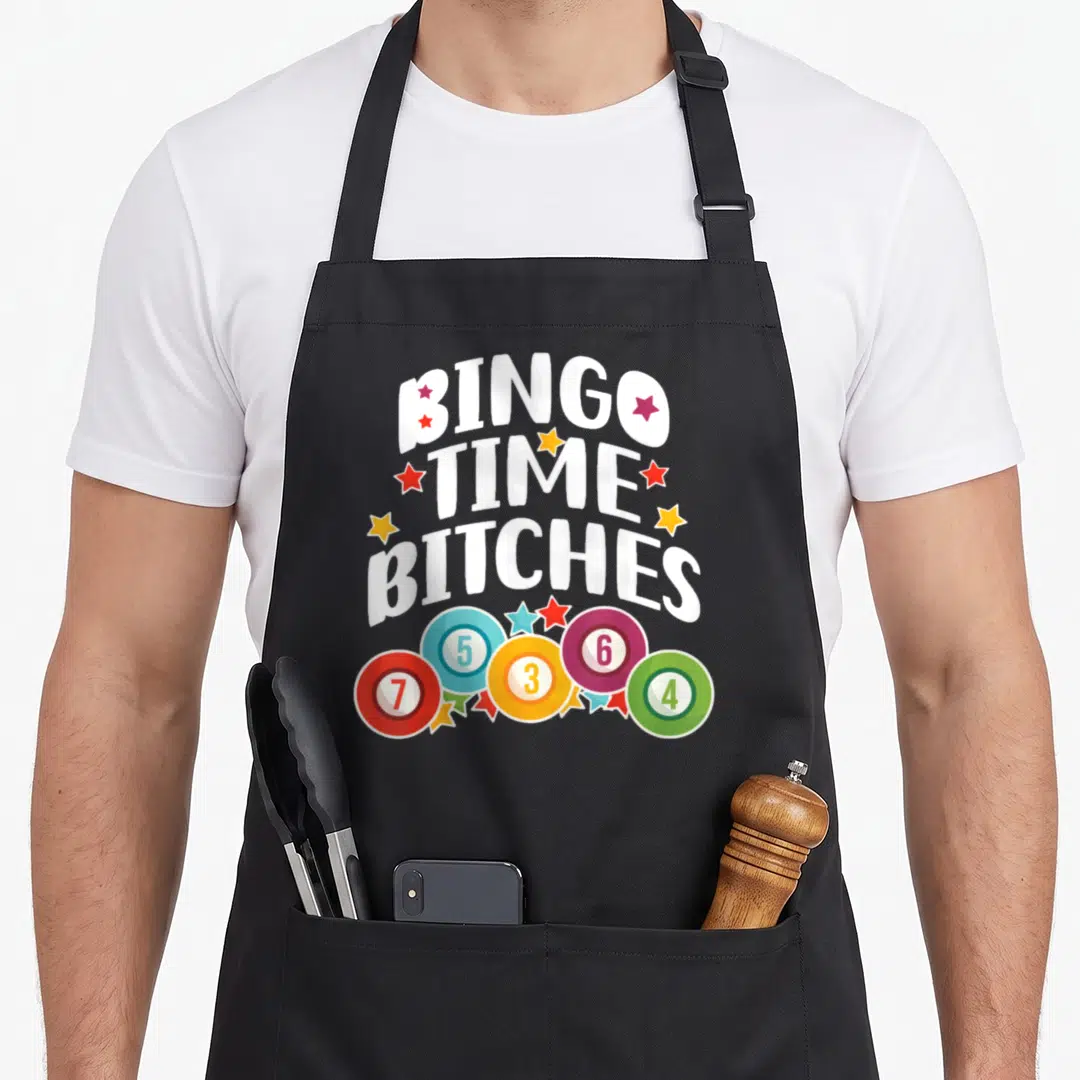 Cute player bingo apron - BINGO TIME BITCHES text surrounded by colorful stars and five numbered bingo chips — shown on a black cotton canvas apron against a white background