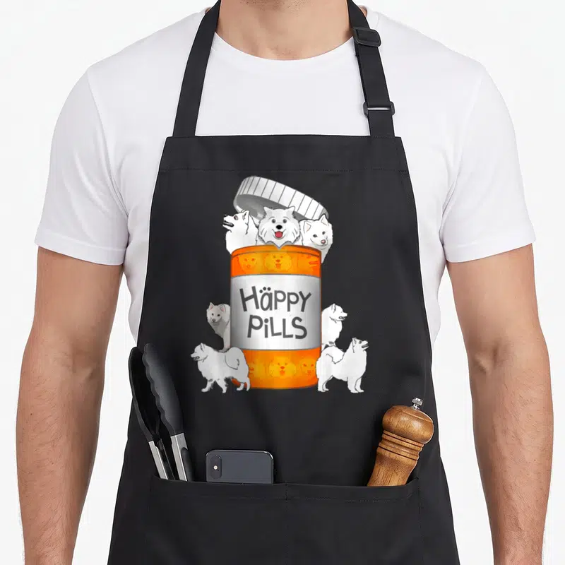 Pill dog apron - Orange pill bottle with six white Samoyed dogs and text Häppy PILLS — shown on a black cotton canvas apron against a white background