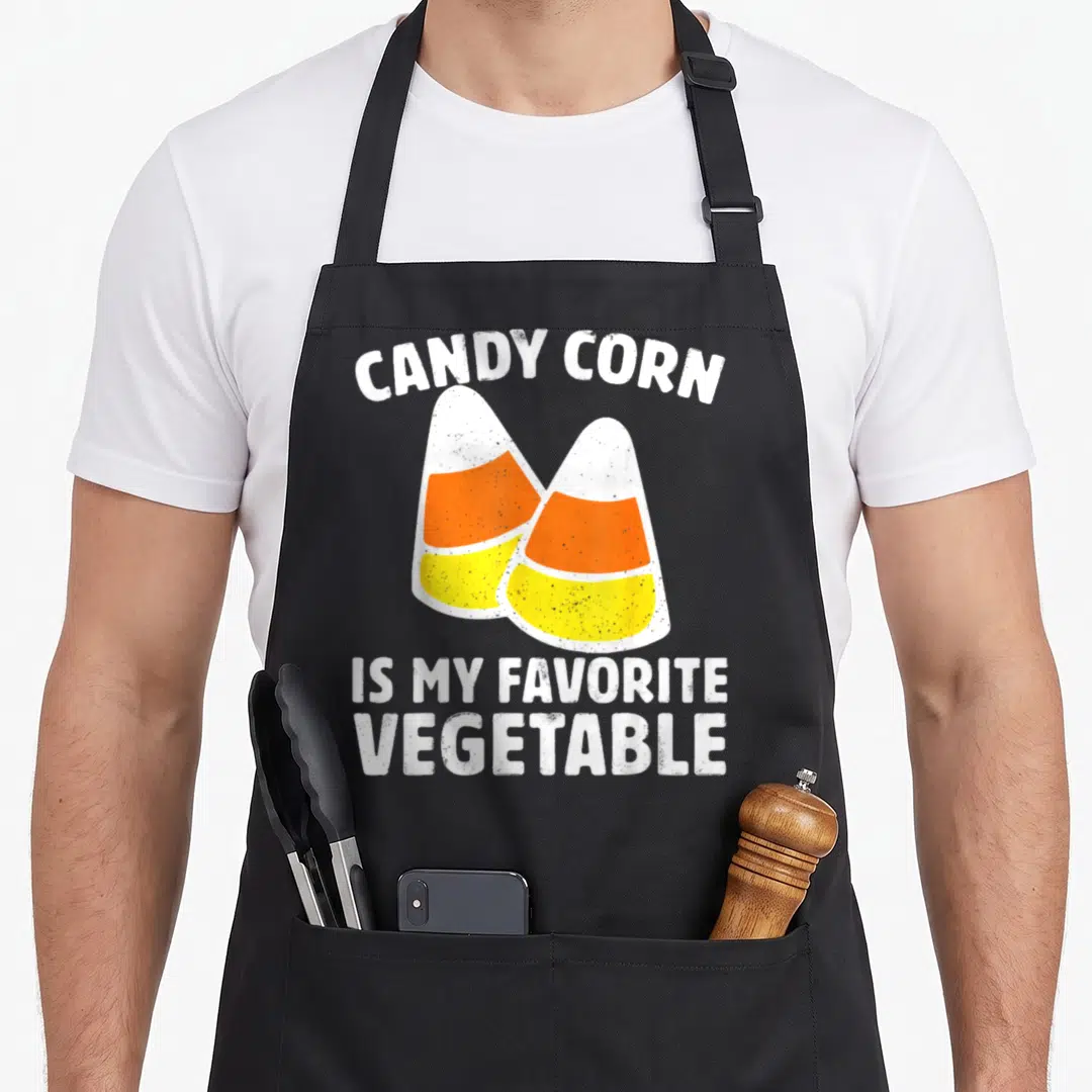 Candy vegetable apron - Two striped candy corns with arched text reading candy corn is my favorite vegetable — shown on a black cotton canvas apron against a white background