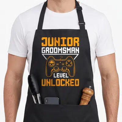 Groomsman gaming junior apron - Orange and white pixel text JUNIOR GROOMSMAN LEVEL UNLOCKED above game controller — shown on a black cotton canvas apron against a white background