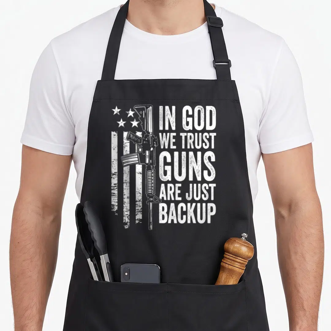 Trust pro apron - White distressed rifle and stars next to stacked text In God We Trust Guns Are Just Backup — shown on a black cotton canvas apron against a white background