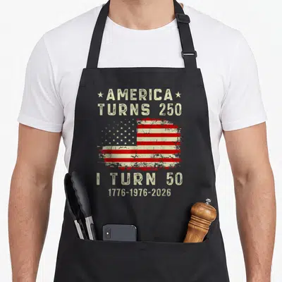50th i turn grills on apron - Distressed US flag with America Turns 250 I Turn 50 text and years 1776-2026 — shown on a black cotton canvas apron against a white background