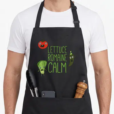 Punny romaine apron - Cartoon tomato lettuce and chili with green LETTUCE ROMAINE CALM text — shown on a black cotton canvas apron against a white background