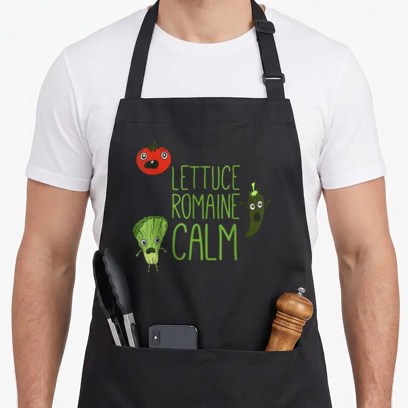 Punny romaine apron - Cartoon tomato lettuce and chili with green LETTUCE ROMAINE CALM text — shown on a black cotton canvas apron against a white background