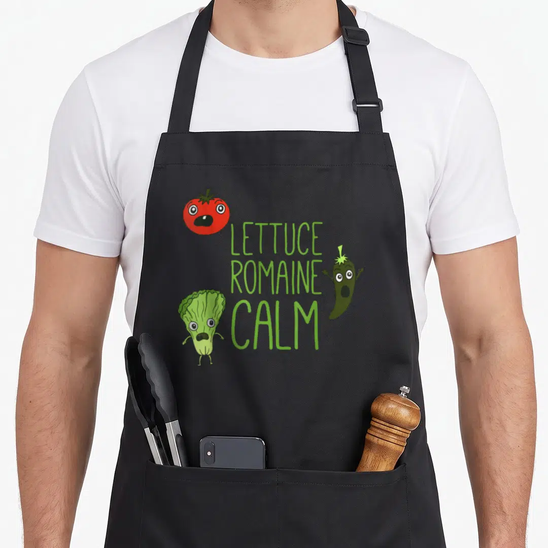 Punny romaine apron - Cartoon tomato lettuce and chili with green LETTUCE ROMAINE CALM text — shown on a black cotton canvas apron against a white background