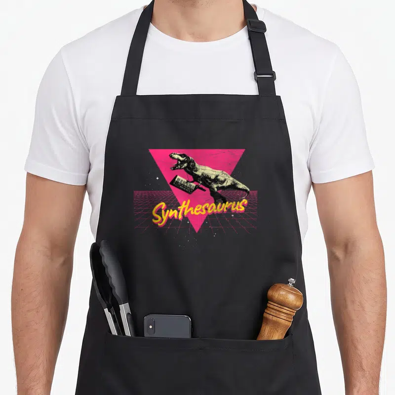 Techno nerd apron - T-Rex holding keytar over pink triangle with Synthesaurus text in yellow script — shown on a black cotton canvas apron against a white background