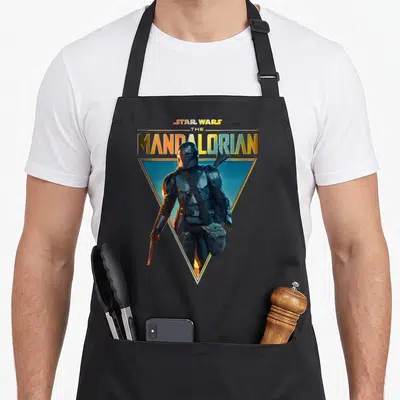Portrait golden apron - Star Wars The Mandalorian text above character photo in gold triangle on blue background — shown on a black cotton canvas apron against a white background