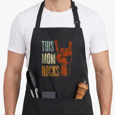 Rock mother's apron - Distressed THIS MOM ROCKS text next to orange rock hand sign — shown on a black cotton canvas apron against a white background
