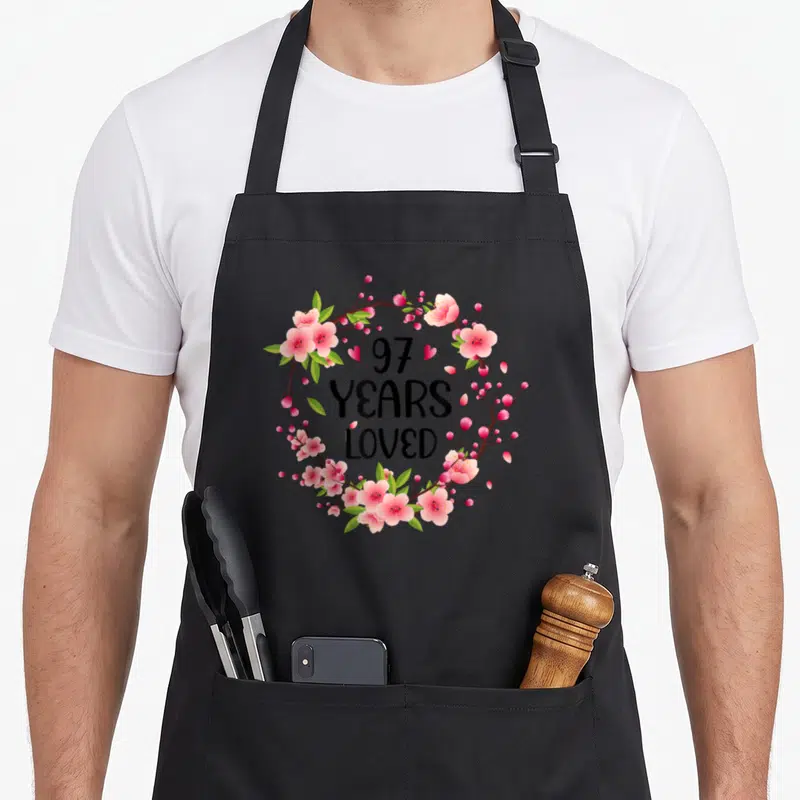 97th floral aprons - Pink floral wreath with 97 YEARS LOVED text and two hearts on black background — shown on a black cotton canvas apron against a white background