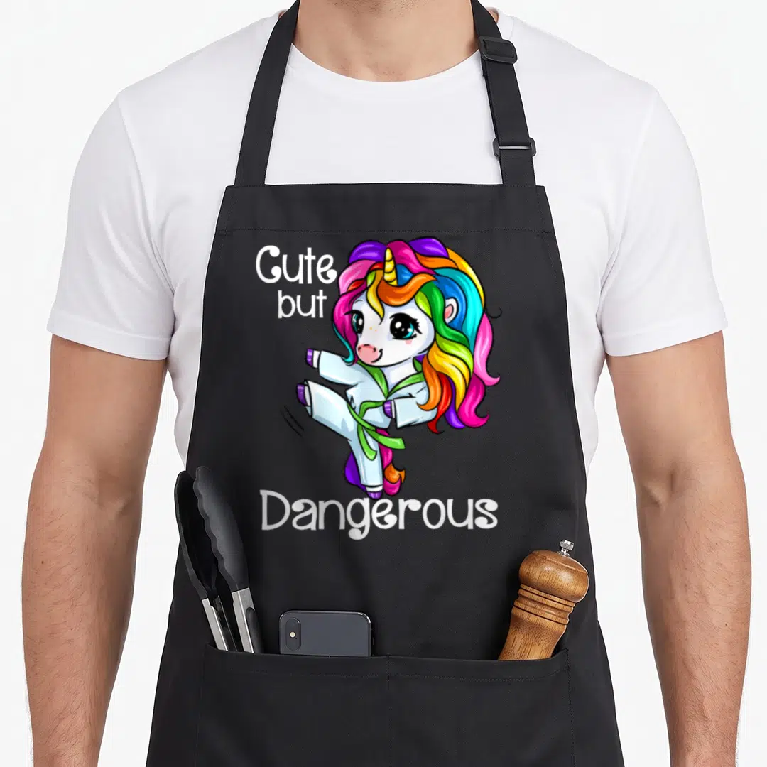 But unicorn apron - Cartoon white unicorn in karate gi with green belt and text Cute but Dangerous — shown on a black cotton canvas apron against a white background