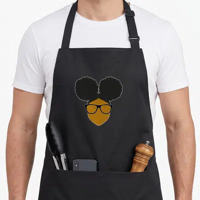 Nerd hair apron - Stylized woman head with two black afro puffs and brown skin wearing black-rimmed glasses — shown on a black cotton canvas apron against a white background