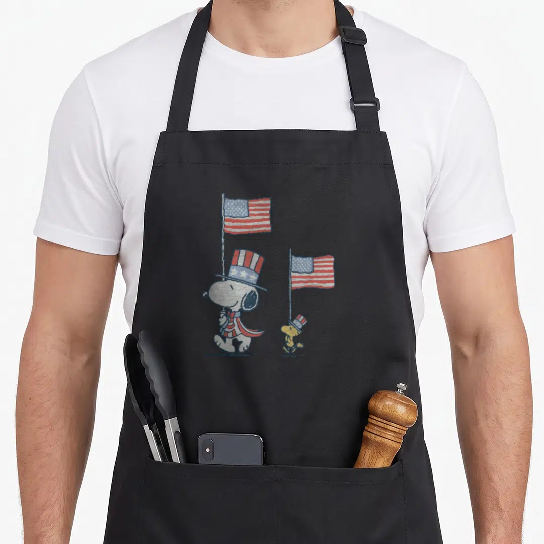 Woodstock independence apron - Snoopy and Woodstock in patriotic top hats holding red white and blue American flags — shown on a black cotton canvas apron against a white background