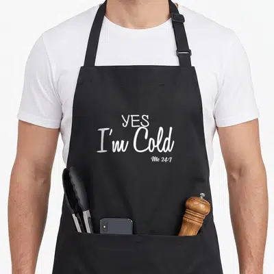 Cold winter humor apron - White text reading YES I'm Cold Me 24:7 stacked vertically — shown on a black cotton canvas apron against a white background