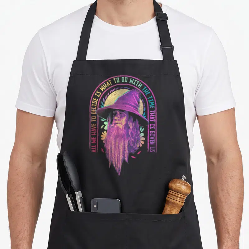 Gandalf ring apron - Retro purple Gandalf portrait inside pink circle with yellow arching quote text — shown on a black cotton canvas apron against a white background