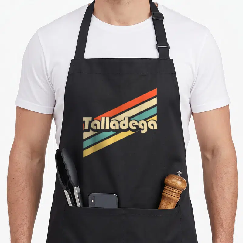 80s alabama apron - Retro Talladega text over four diagonal stripes in orange cream teal and yellow — shown on a black cotton canvas apron against a white background
